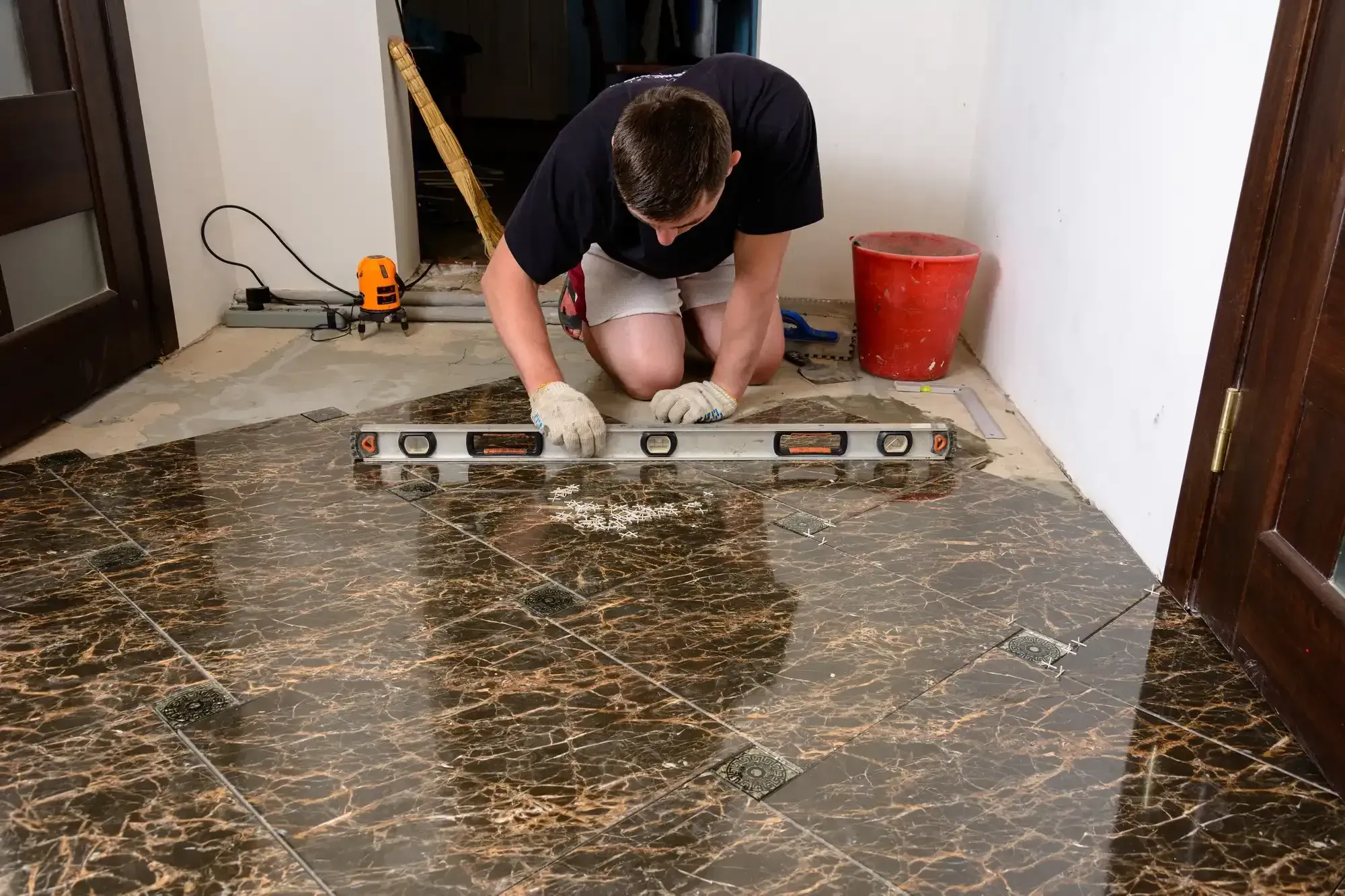 Person kneeling, using level on brown and black tile floor, near doorway.