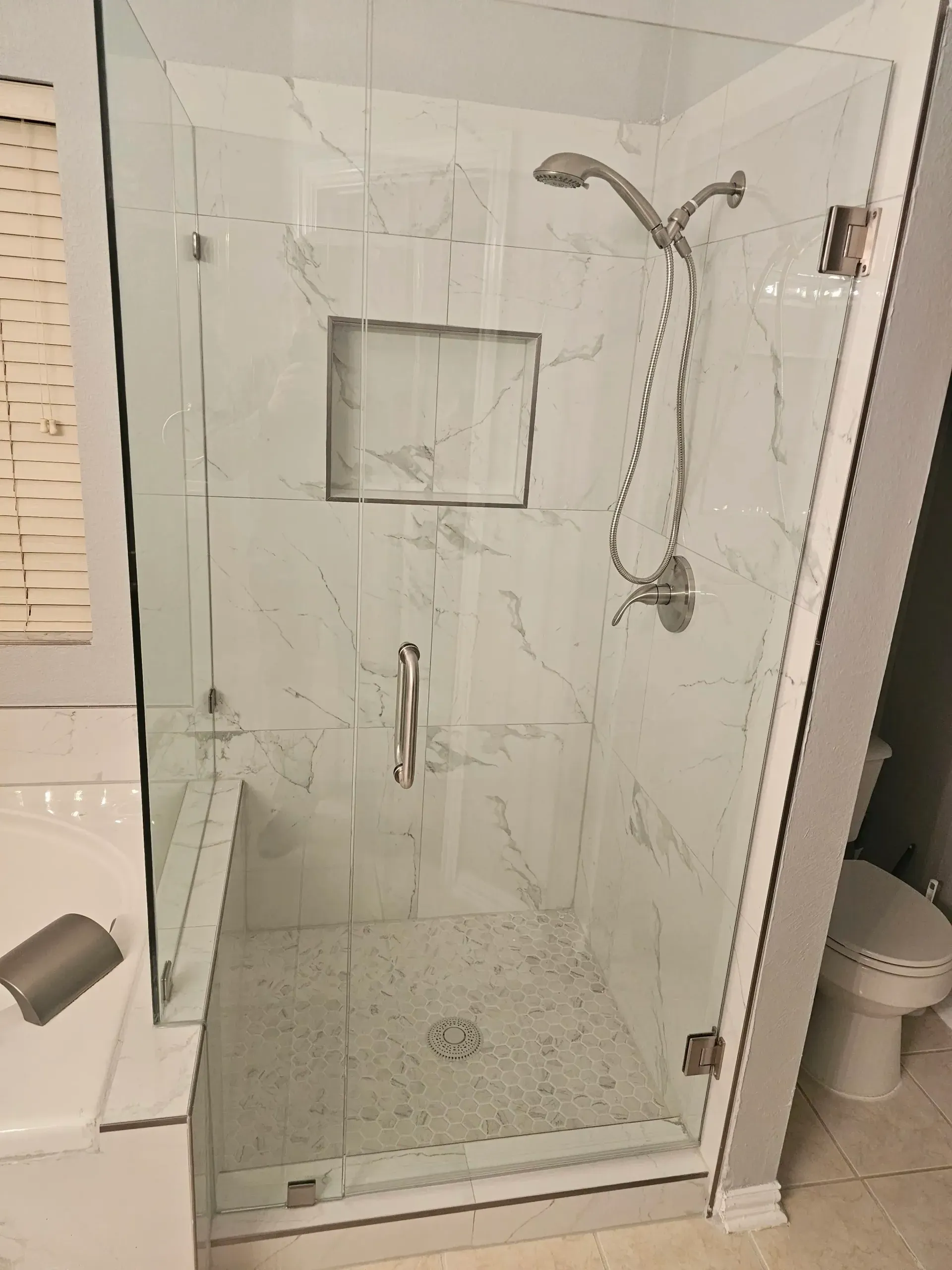 Glass shower enclosure with white marble-look tiles, a built-in shelf, and a shower head.