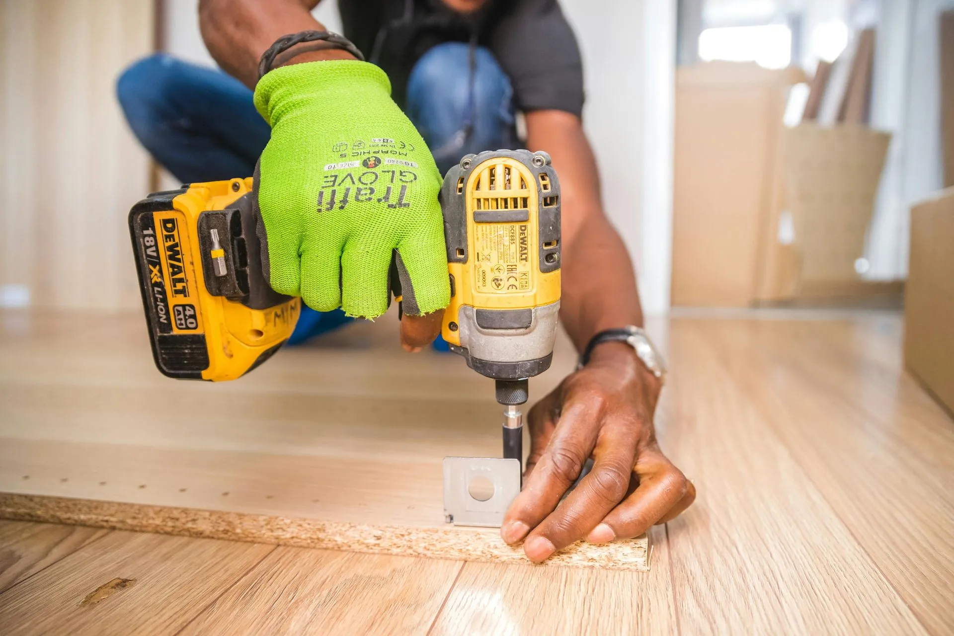 Person using a power drill on a wooden floor, wearing a green glove.