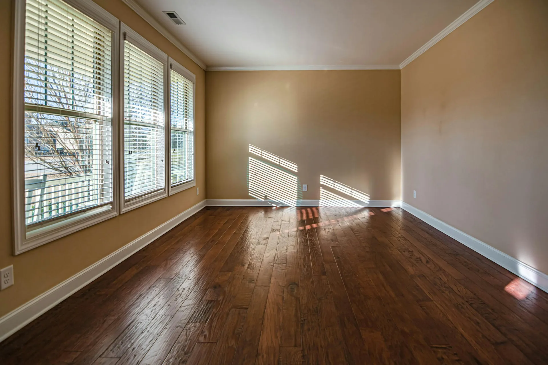 Empty beige room with hardwood floors and large windows casting sunlight on the wall and floor