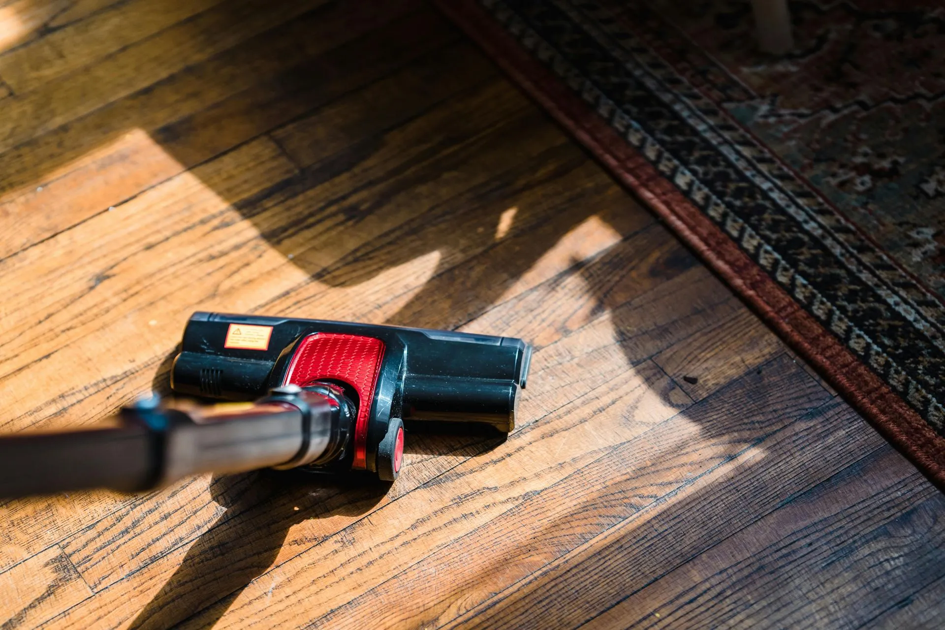 Vacuum cleaner head on a sunlit hardwood floor next to a patterned rug